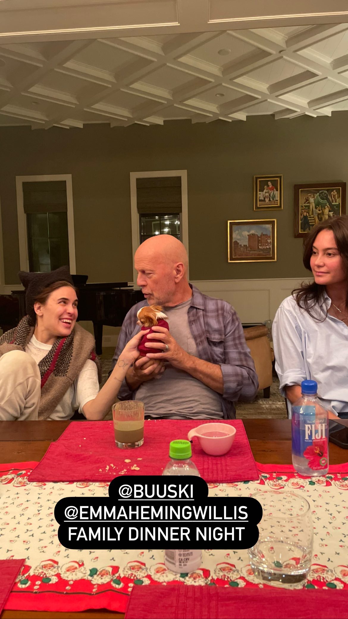 Bruce Willis sentado à mesa com a filha e esposa enquanto segura cachorro com as mãos.