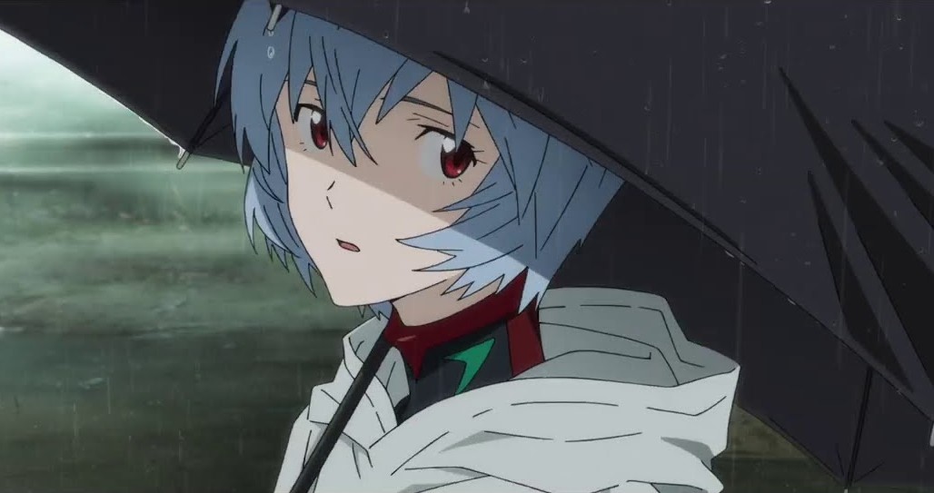Audiodescrição resumida: Rei Ayanami, menina branca, de olhos vermelhos e cabelos azuis. Está com a boca aberta, olhando para a esquerda enquanto segura um guarda-chuva. Ela usa um moletom branco.